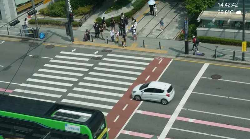 Live view of a busy crosswalk in Seoul on a summer afternoon, with time and temperature on screen